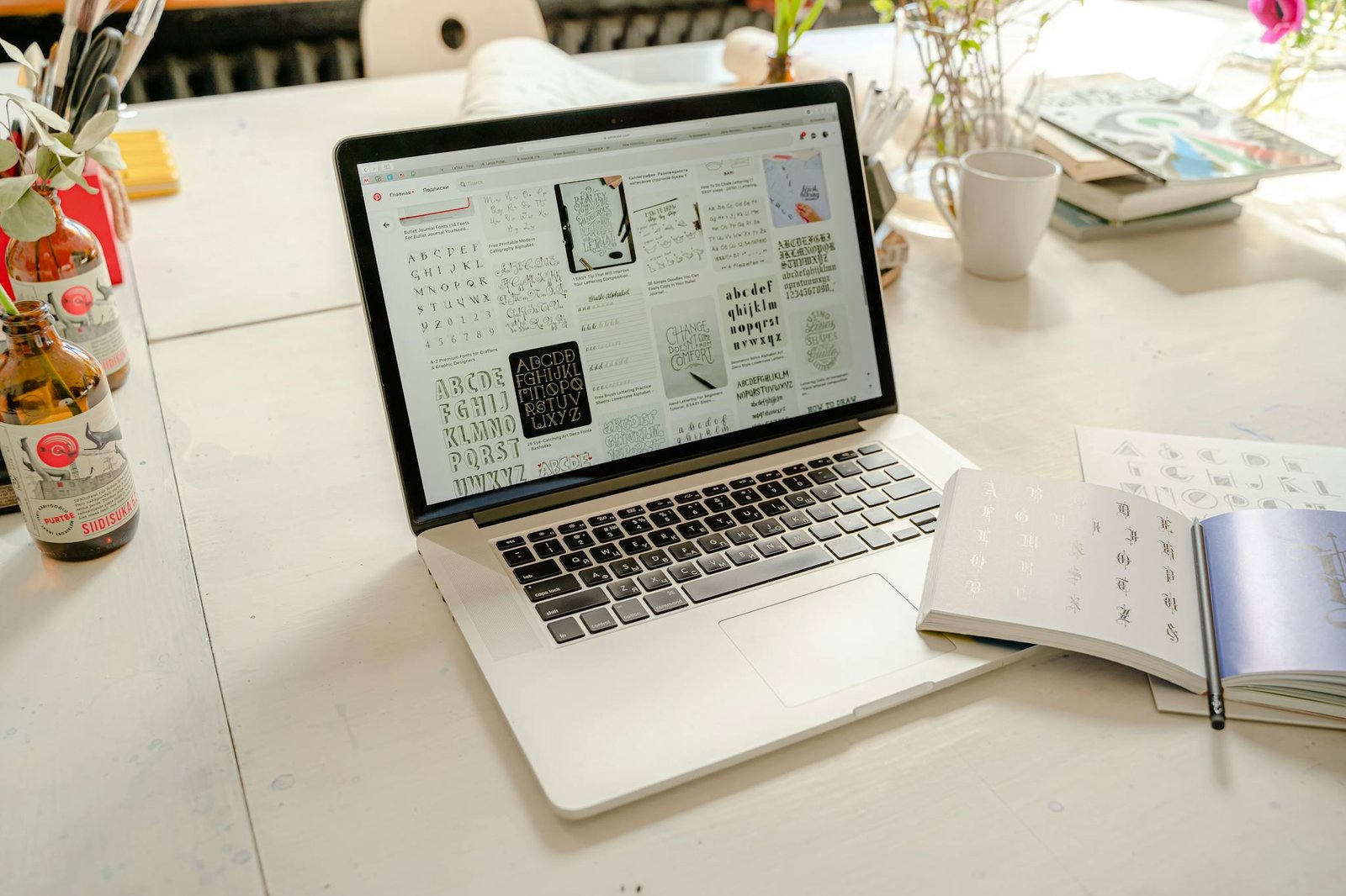 Bright creative workspace featuring laptop with calligraphy designs, books, and artistic tools.